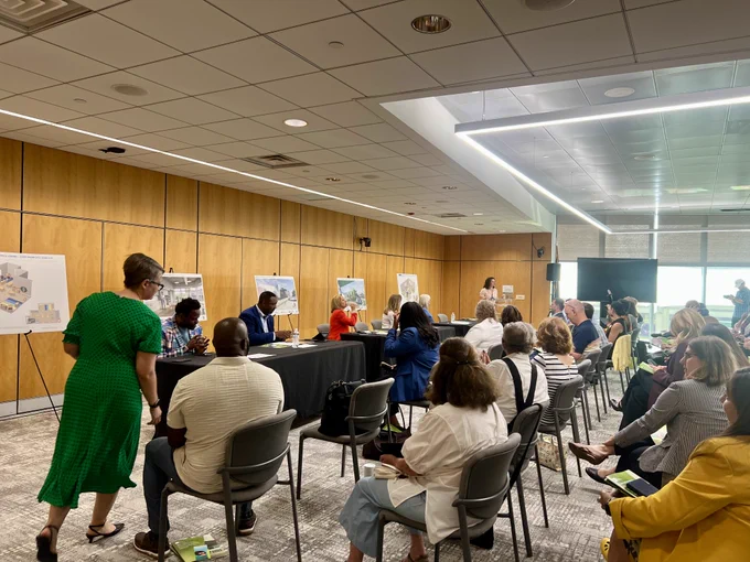 A conference room with people seated and a panel of speakers at the front. The room has wooden walls with presentation boards displayed. Attendees are listening attentively, and a person is walking towards the panel. The environment is professional and focused.
