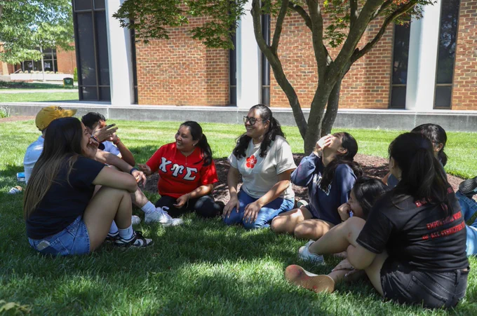A group of college students sit in a loose circle on the grass under a leafy tree beside a brick academic building with large windows. They’re talking and laughing together in the shade. One student wears a red shirt with Greek letters “ΧΥΣ.” The scene feels relaxed and social, capturing a casual moment of community on campus.
