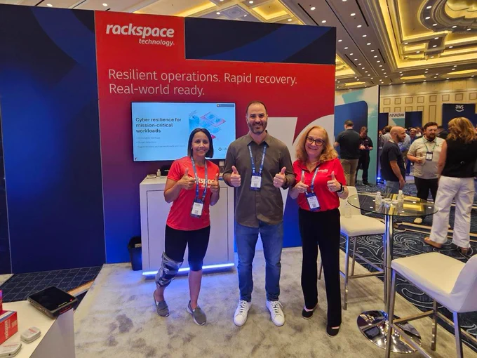 A group of three people standing in front of a Rackspace Technology booth at a conference. Two are wearing matching red Rackspace shirts, and one person is in a gray shirt. They are all smiling and giving thumbs up. The backdrop features a Rackspace logo and promotional text. Other conference attendees are visible in the background.
