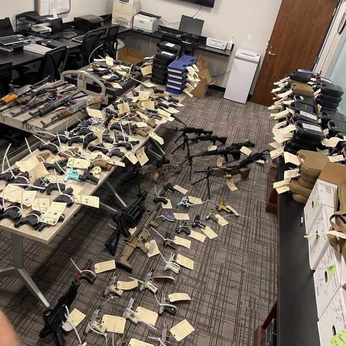 Room filled with numerous firearms laid out on tables and floor, each tagged with identification labels, resembling evidence storage.