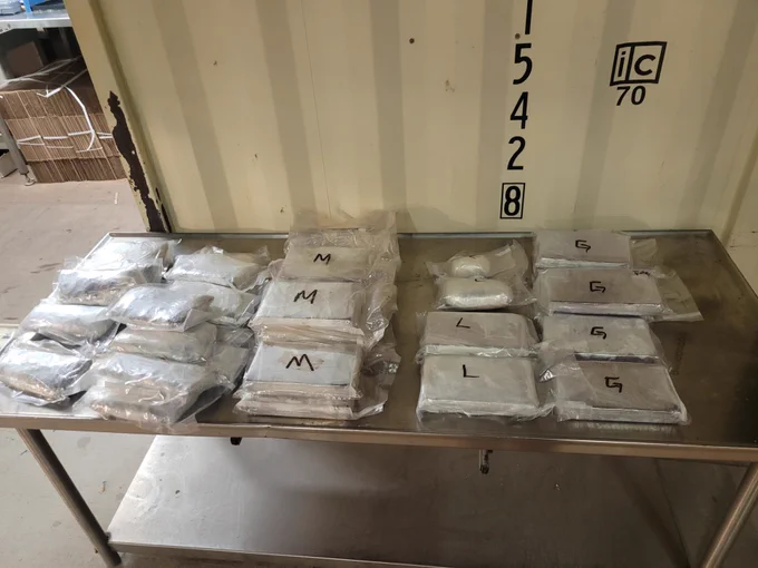 Multiple clear plastic bags filled with narcotics, labeled with letters M, L, and G, arranged neatly on a metal table in an industrial setting.