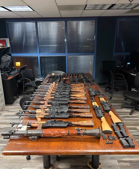 A large collection of various rifles and firearms neatly arranged on a wooden table inside an office setting.