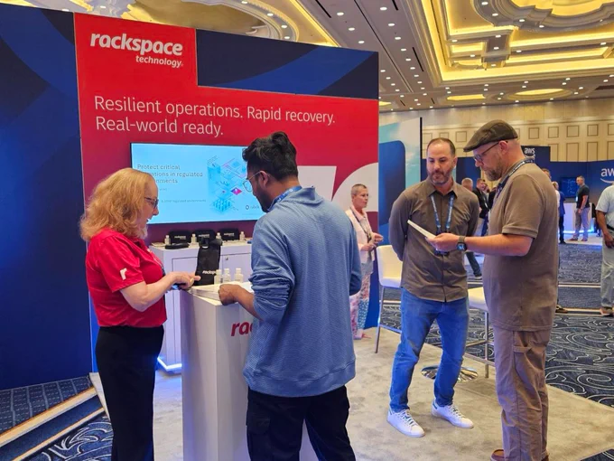 A group of people are gathered at a Rackspace Technology booth in a convention center. The booth displays text promoting resilient operations and rapid recovery. Attendees are engaged in conversation and examining materials. The setting features elegant lighting and a carpeted floor.