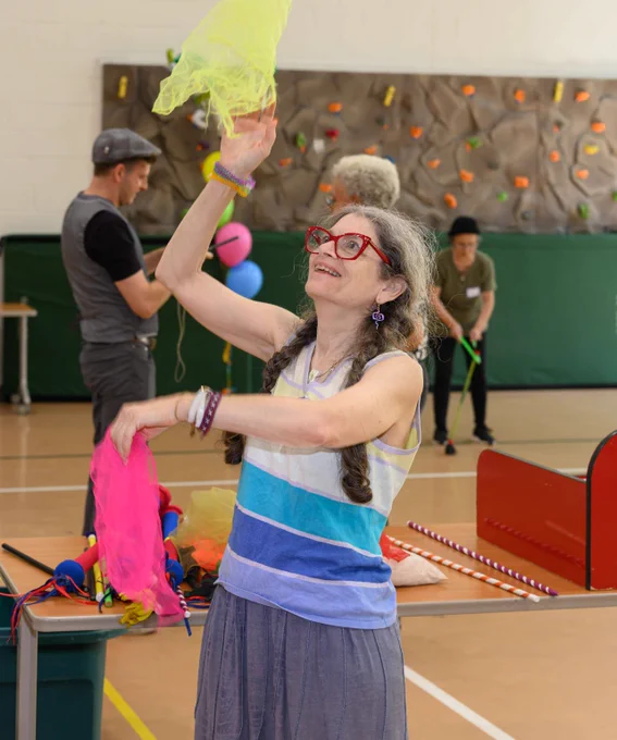Woman juggling colorful scarves in a gymnasium with a rock climbing wall and other people in the background.