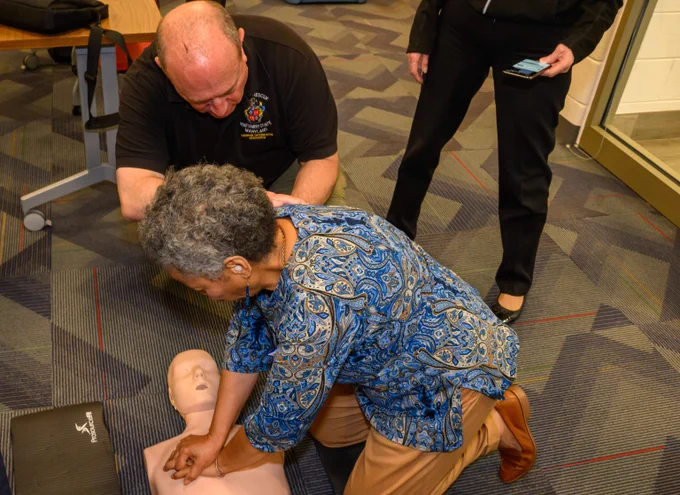 An elderly woman practices chest compressions on a CPR manikin under the guidance of an instructor in a training room.