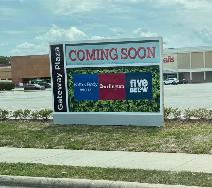 A sign announcing the upcoming opening of stores at Gateway Plaza, including Bath & Body Works, Burlington, and Five Below. The sign is located in a mostly empty parking lot under a cloudy sky.