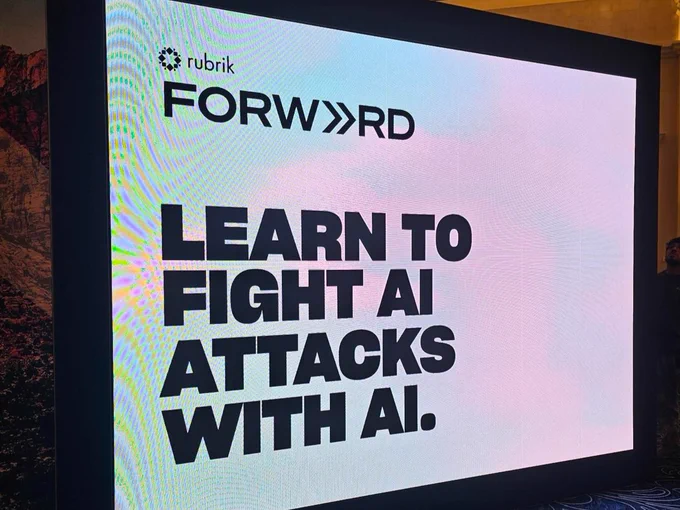 The image shows a digital display with the Rubrik logo and the word "FORWARD." Below, the text reads, "LEARN TO FIGHT AI ATTACKS WITH AI." The background is a gradient of light colors.