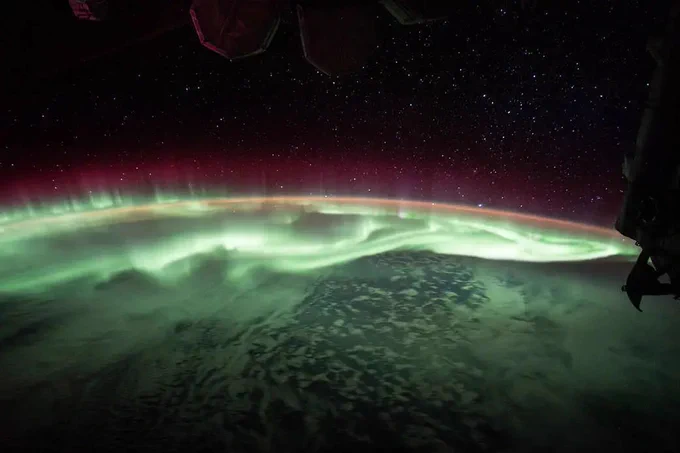 A photograph taken from the International Space Station, parts of which can be seen at the top and right of the image, showing a massive, stunning green and red aurora on Earth.