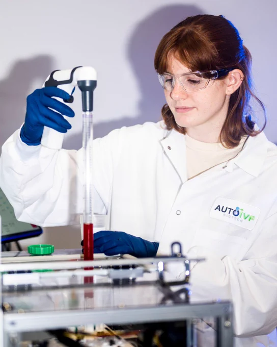 Young female scientist with reddish-brown hair and safety glasses, wearing a white lab coat with \"AUTO IVF BIOTECH\" logo and blue gloves, meticulously pipetting a red liquid in a lab setting. Lab equipment is visible in the foreground.