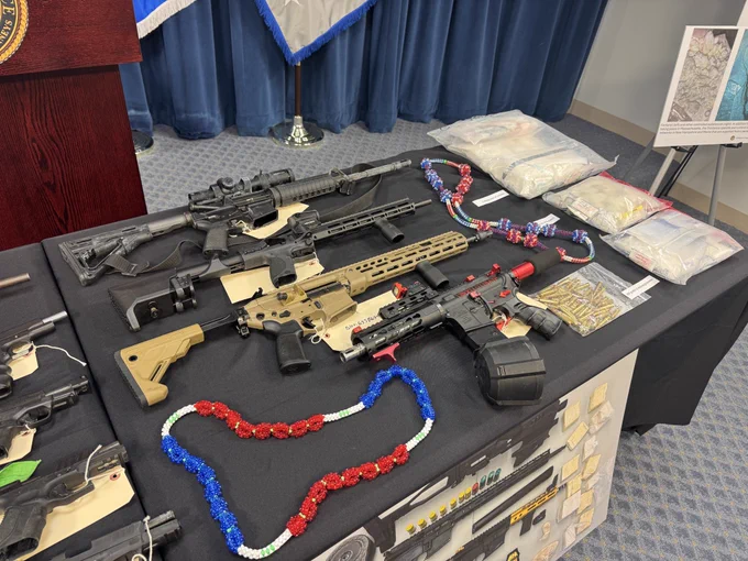 Close-up of evidence seized during a federal gang investigation, including several rifles, ammunition, suspected narcotics, and red, white and blue bead necklaces associated with the Trinitarios. The items are displayed on a table before a press conference at the U.S. Attorney’s Office in Boston.