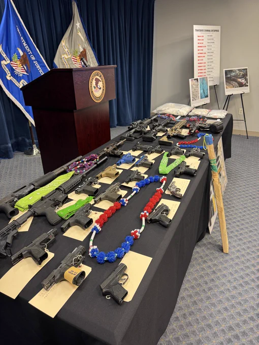 Evidence displayed ahead of a federal press conference announcing charges against alleged Trinitarios gang members. Multiple firearms, including rifles and handguns, ammunition, narcotics, and gang-affiliated bead necklaces are arranged on a table beside a DOJ podium and flags at the U.S. Attorney’s Office in Boston.