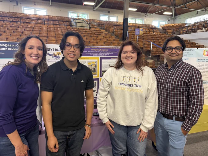 Group of four people standing in front of display boards at an event. One display features "Tennessee Tech" branding. The group is in a large hall with wooden bleachers in the background.