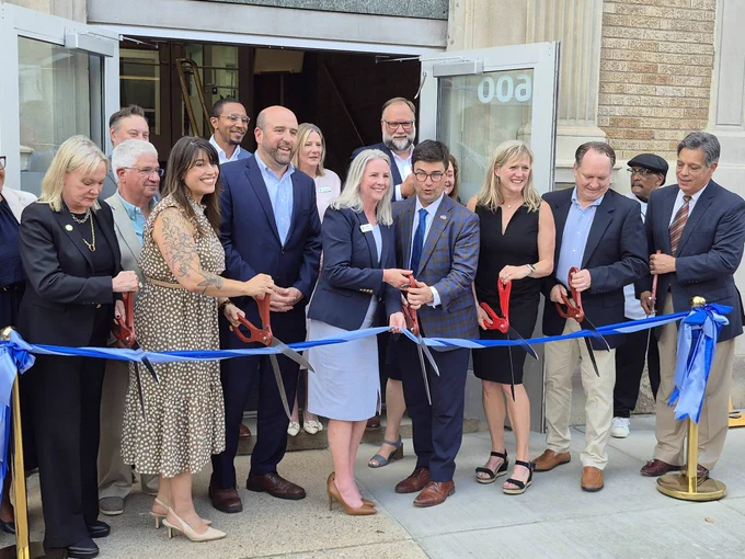 Secretary Rick Siger and local officials standing behind a blue ribbon, holding large scissors for a ribbon-cutting ceremony outside the YMCA entrance.