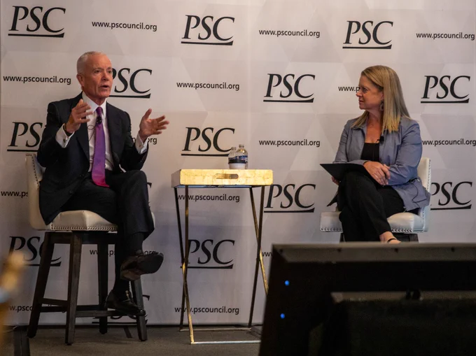 Two professionals engaged in a discussion on stage at a PSC event, seated with a small table and water bottle between them.