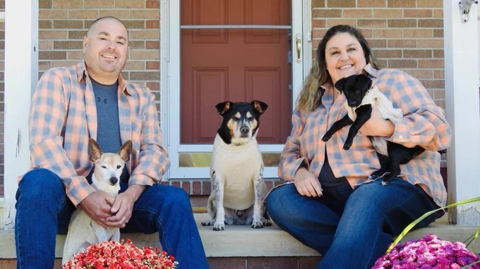 A couple wearing matching plaid shirts and jeans poses on the front steps of a brick house with three dogs.
