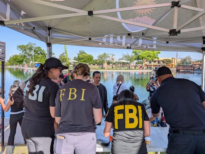 FBI agents in black shirts with yellow letters working under a canopy by a lakeside with people gathered nearby.