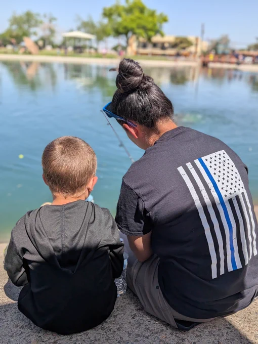 An FBI agent wearing a thin blue line t-shirt and a young boy sit side by side by a calm lake, fishing; both facing the water on a sunny day.