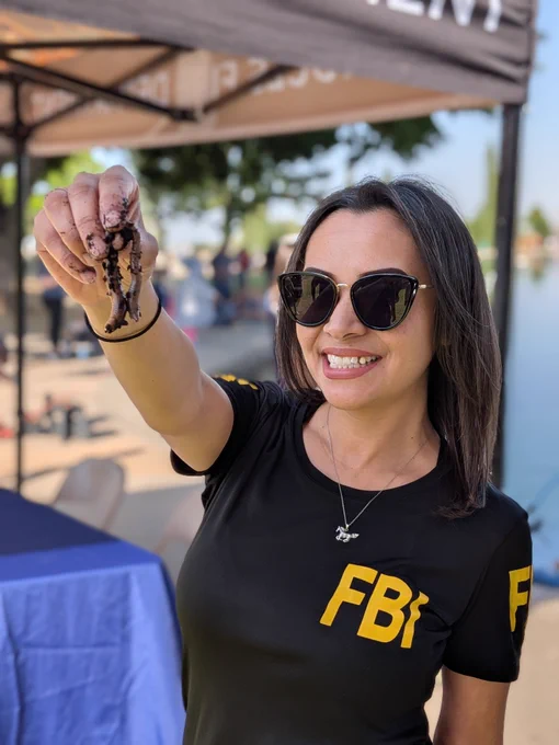 Person wearing a black FBI shirt holding up a worm near a waterfront event tent.