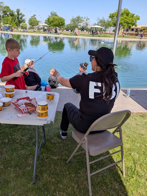 Woman in FBI shirt sitting on a chair by a lake, helping children with fishing bait, fishing on the water's edge under a sunny sky.