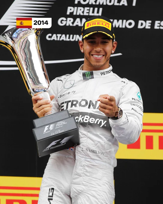 Lewis Hamilton on the podium holding the trophy after winning the 2014 Spanish Grand Prix. He is wearing his white race suit, and smiling while pointing at the camera with his left hand.