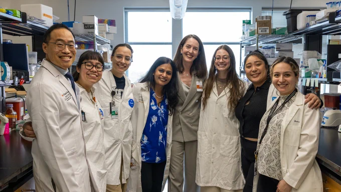 Eight people stand together in a brightly lit laboratory. Shelves behind them hold boxes, bottles, pipettes, and other lab supplies. Most of the group members wear white lab coats, and some have ID badges. The space features large windows in the background of an active research lab.