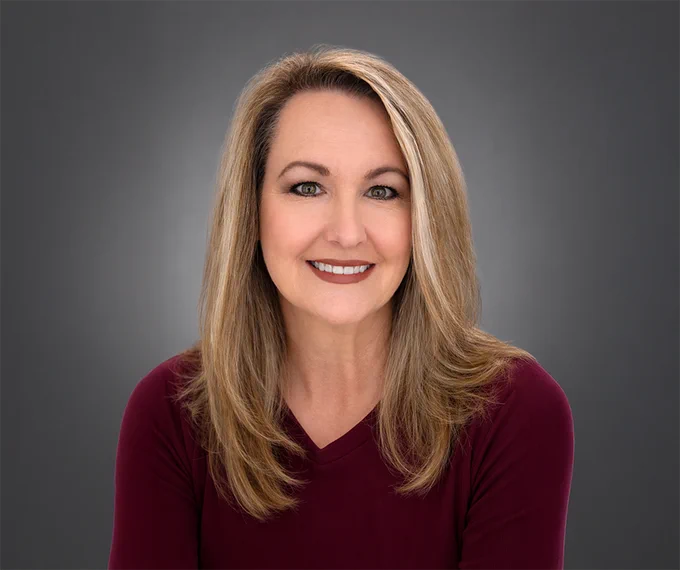 A professional headshot of a smiling woman with long hair against a grey background. The image conveys a friendly and approachable mood.