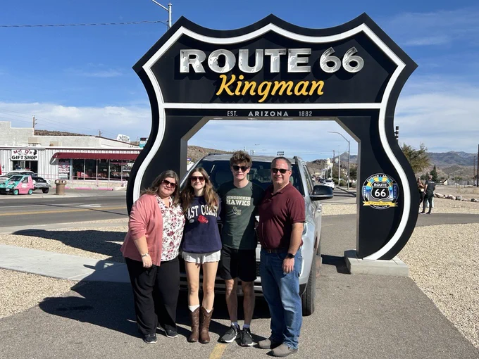 Four people posing under the large Route 66 Kingman sign on a sunny day with a clear blue sky and desert landscape.