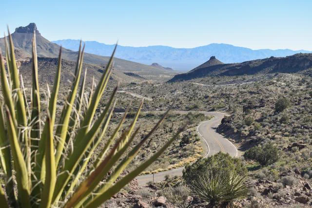 Winding road cutting through a dry, rocky desert landscape with distant mountains under a clear blue sky.