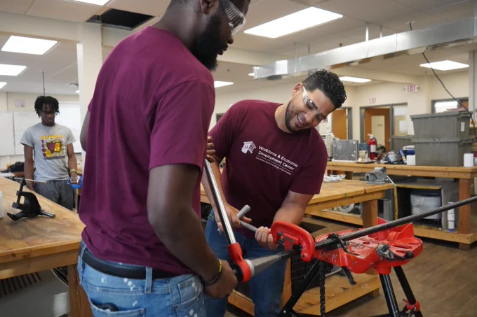 Two students in a workshop using a pipe wrench to tighten a metal pipe secured in a red pipe vise on a workbench.
