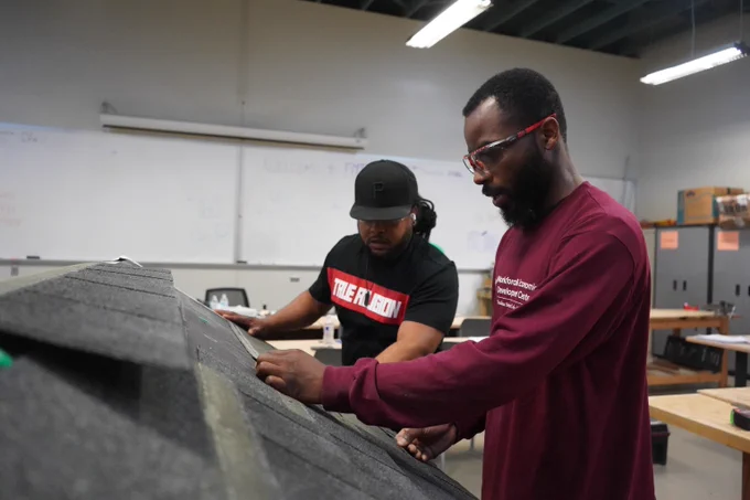 Two individuals examine a roofing model indoors, focusing on the shingles and structure in a workshop setting.