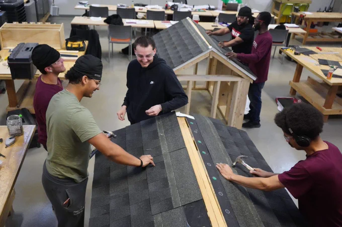 Group of students working together on constructing and shingling small wooden roof models in a workshop classroom.