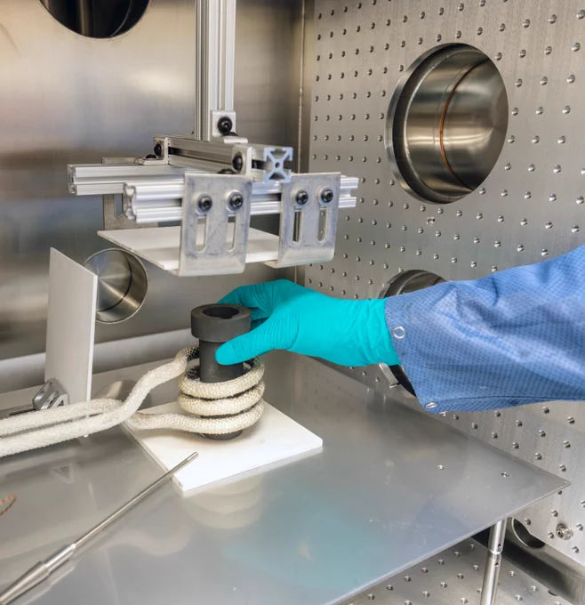 Gloved hand placing a black cylindrical object wrapped with white rope inside a stainless steel testing chamber.