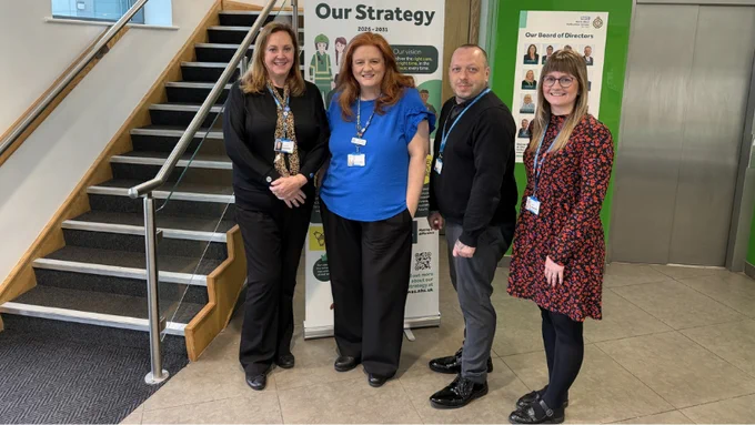 A group of four people stand in an indoor setting near a staircase. Behind them is a banner displaying "Our Strategy". The individuals are wearing name badges and are smiling for the photo.