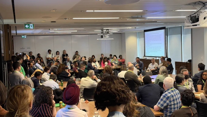  A wide view of attendees gathered at the A11yVerse x IAAP Canada Summit in a conference space at Deloitte in Toronto. Participants are seated around tables and standing along the walls while listening to a panel discussion taking place at the front of the room. A large presentation screen displays discussion notes, and attendees wearing conference badges engage in conversations about accessibility, innovation, and leadership. The room is filled with accessibility professionals, entrepreneurs, advocates, and ecosystem leaders participating in the event. 
