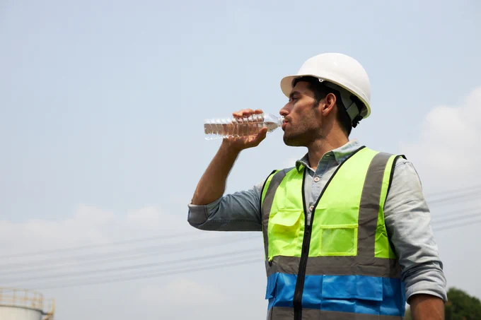 a person wearing a hard hat and safety vest is taking a drink from a bottle of water.