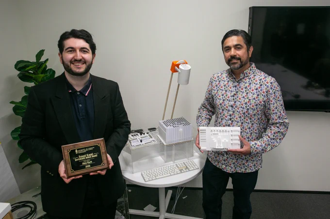 Alex Kurk stands on the left, holding a wooden award plaque engraved with the Troost ILead Difference Maker Award. Sanjay Malaviya stands on the right, holding a white 3D-printed model of a software interface. Between them is a round table with several 3D-printed models, a keyboard and a clear enclosure. 