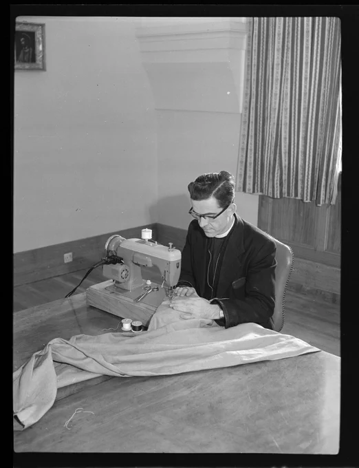 Black and white image of a bespectacled man sitting at a table with a sewing machine and large piece of fabric. He is wearing a dark suit with clerical collar. The room appears fairly bare behind him with one framed image on the wall and curtains at window.