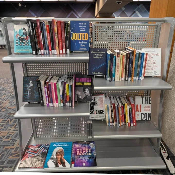 A metal book shelf with books. Some of the covers face outwards, and some of the spines face outwards.