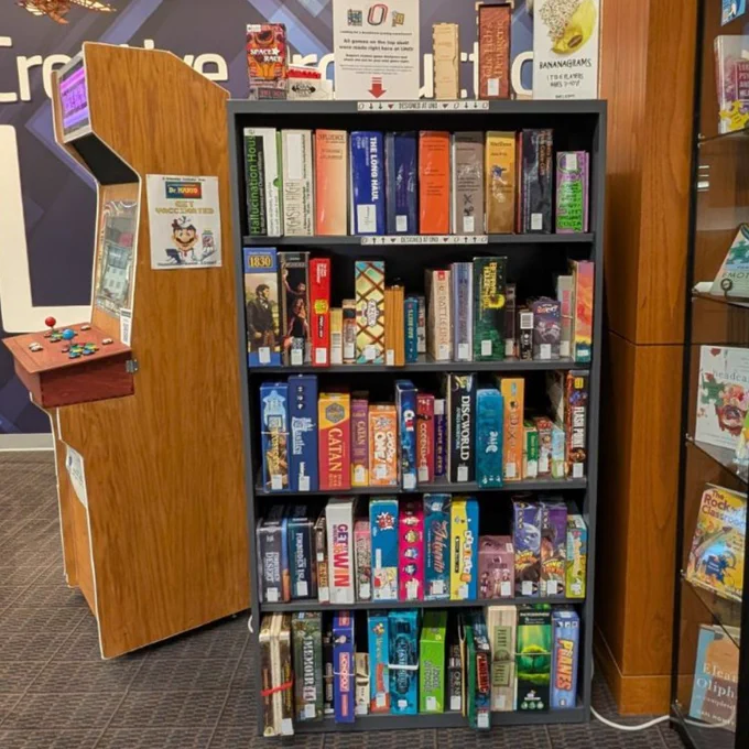 A metal shelf with board games on it. There is a wooden arcade machine next to the metal shelf.