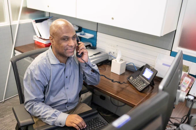 A blind professional sits at a desk, answering a phone call while typing on a desktop computer.