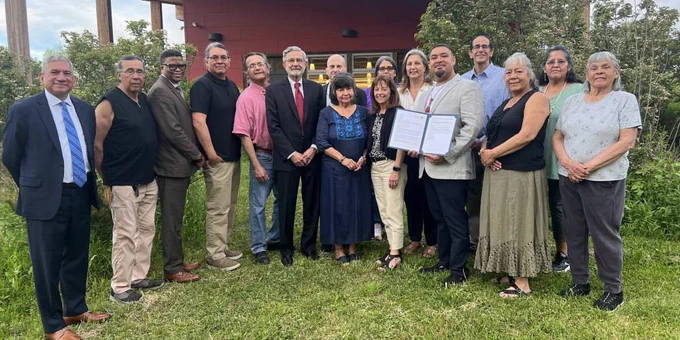 A group of people is standing outdoors in front of a building, with greenery surrounding them. One individual is holding a framed document. Everyone is dressed in a mix of formal and casual attire.