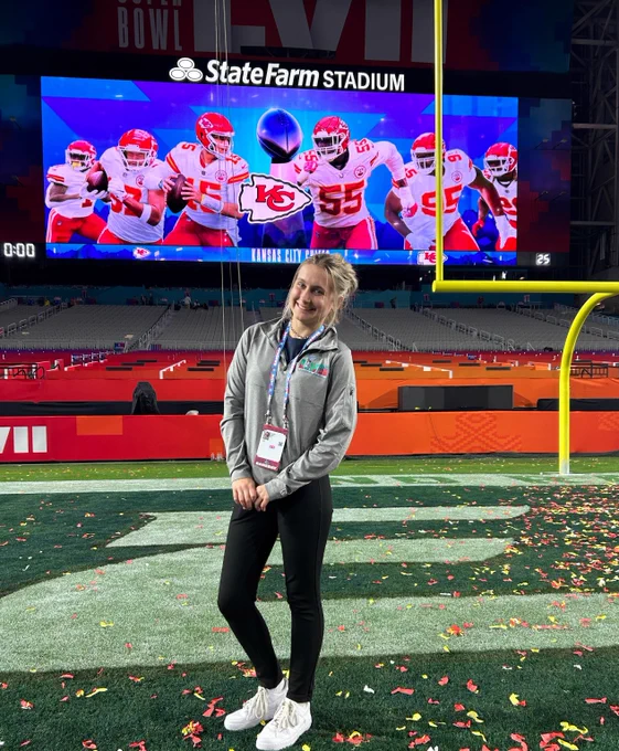 A person is smiling and standing on a football field for the Super Bowl covered with scattered confetti. In the background, a large video screen displays images of Kansas City Chiefs players with the State Farm Stadium logo above.
