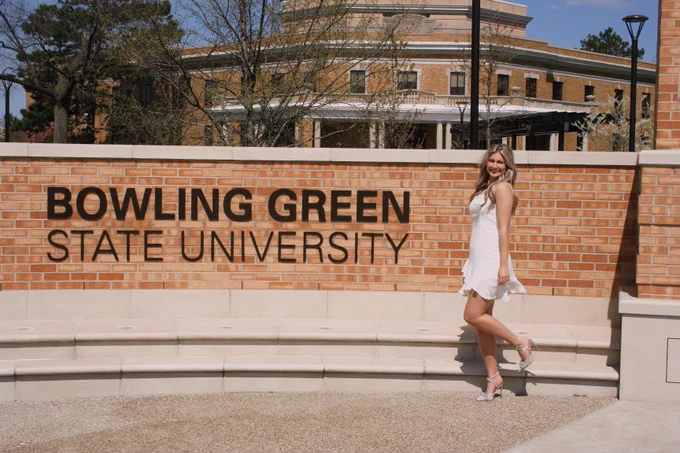 A person in a white dress is standing and smiling next to a brick wall with the text "Bowling Green State University." A building and trees are visible in the background.
