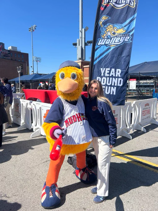 A person stands next to a colorful baseball mascot wearing a striped jersey and cap, holding a baseball bat. There's a Toledo Walleye banner in the background with the text "Year Round Fun". They appear to be at an outdoor event near a stadium.