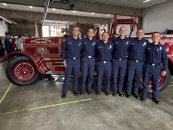 Six members of the Huntington Beach Fire Department's 2026 Firefighter Paramedic Academy pose in front of the department's historic antique fire engine during their graduation ceremony. The graduates, dressed in navy blue uniforms, stand shoulder to shoulder inside a fire station apparatus bay as they celebrate the completion of their academy training.