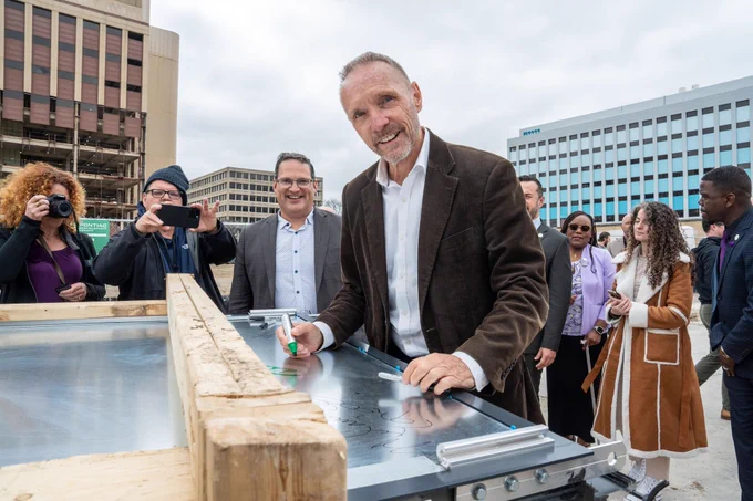 Oakland County Executive Dave Coulter leaning over a metal sheet to sign it as onlookers stand nearby in Downtown Pontiac.