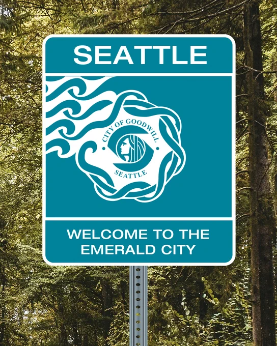 A road sign with "Seattle" at the top, featuring the city of Seattle design with waves and the "City of Goodwill" emblem. Below it reads, "Welcome to the Emerald City." The background shows green trees.
