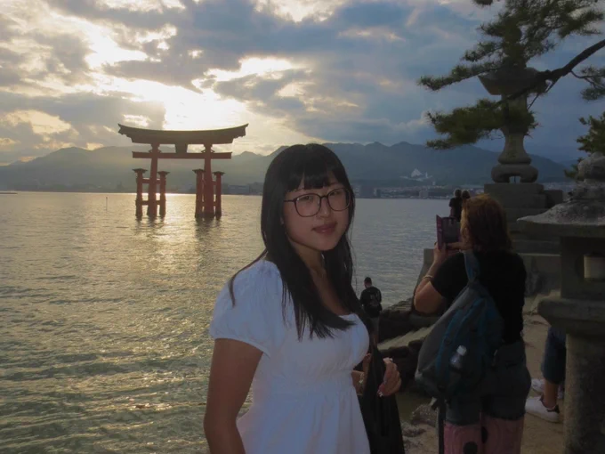 A person stands by the water at sunset, with Itsukushima Shrine's torii gate visible in the background. Other visitors and trees are in the scene, under a partly cloudy sky.