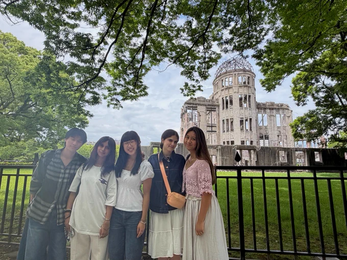 A group of five people standing in front of the Hiroshima Peace Memorial, also known as the Atomic Bomb Dome, surrounded by lush greenery.