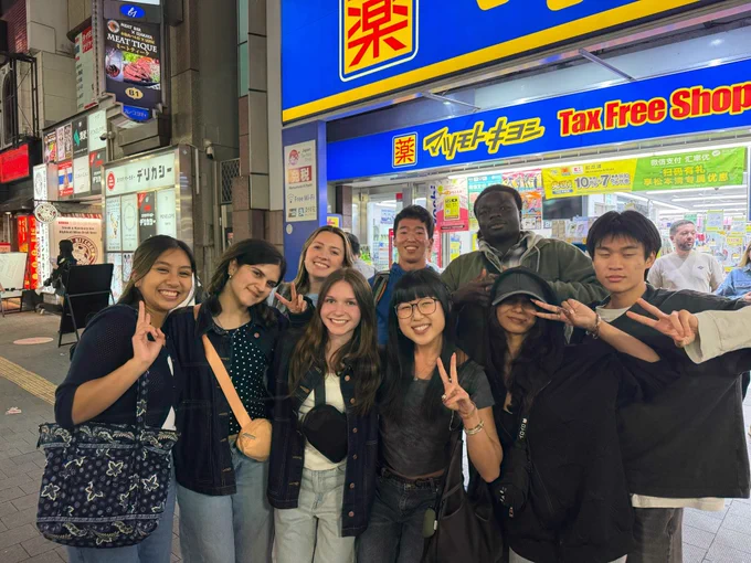 A group of people smiling and posing in front of a brightly lit store with a "Tax Free Shop" sign in Japan. The setting appears to be a busy urban street at night.